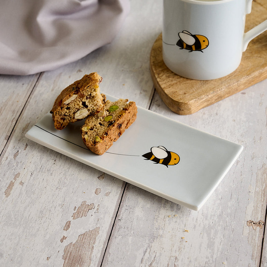 A light grey rectangular fine bone china mini tray with Bee by Jin Designs, styled on a rustic white wooden table next to a matching light grey mug on a wooden coaster.
