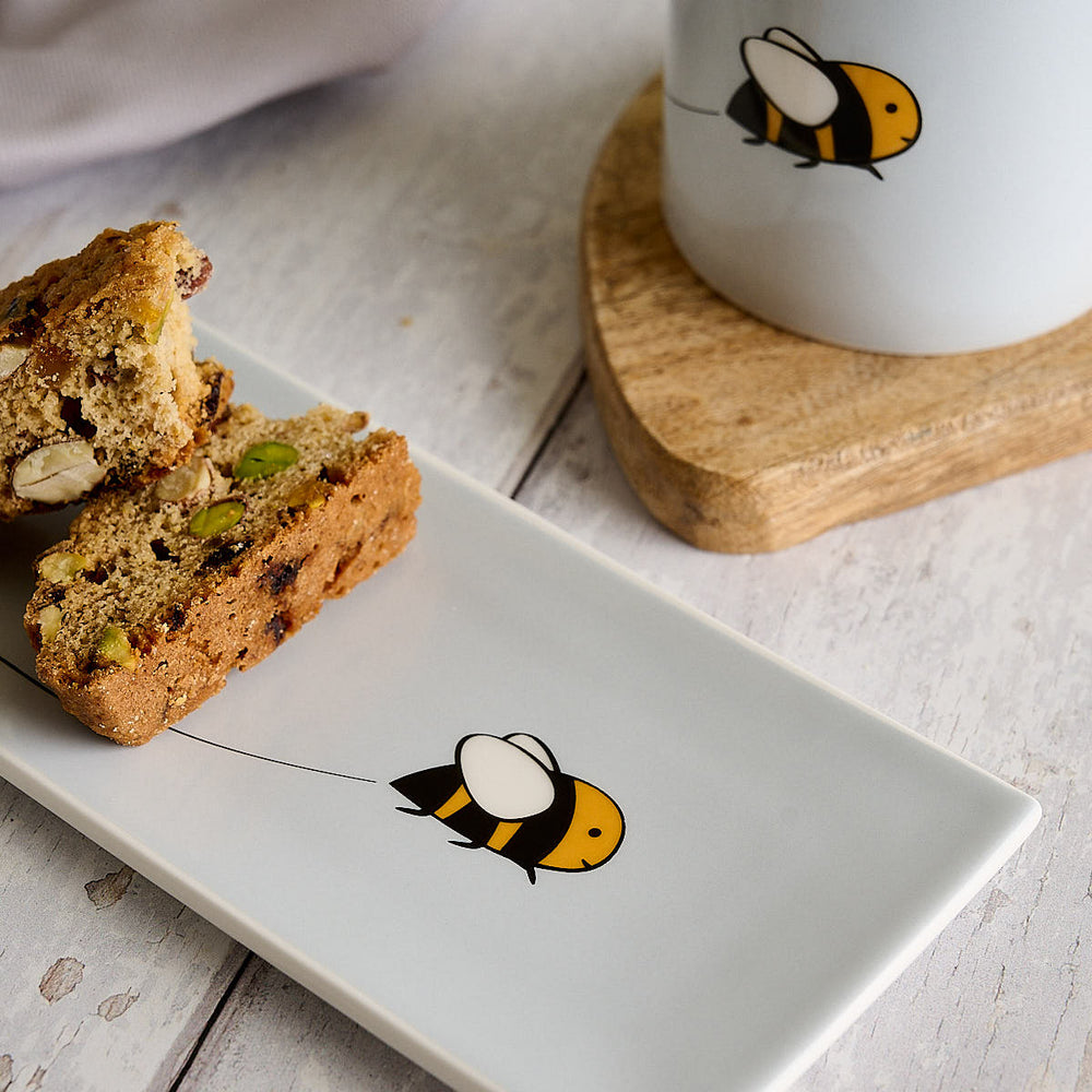 A light grey rectangular fine bone china mini tray with Bee by Jin Designs, styled on a rustic white wooden table next to a matching light grey mug on a wooden coaster.