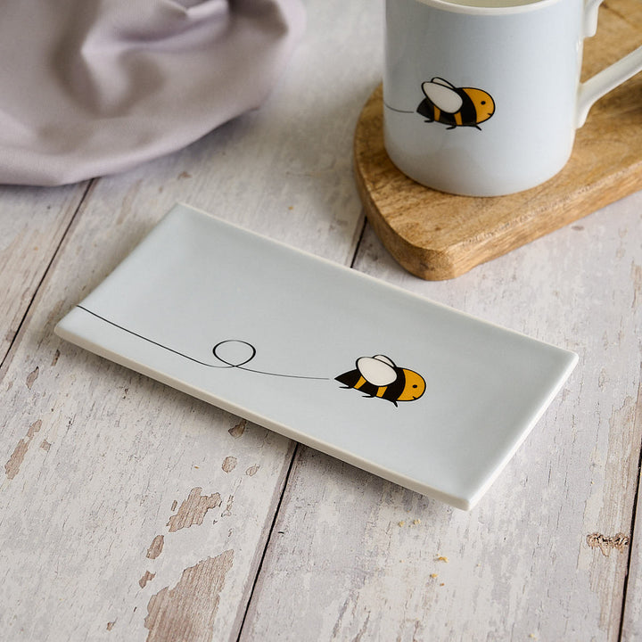 A light grey rectangular fine bone china mini tray with Bee by Jin Designs, styled on a rustic white wooden table next to a matching light grey mug on a wooden coaster.