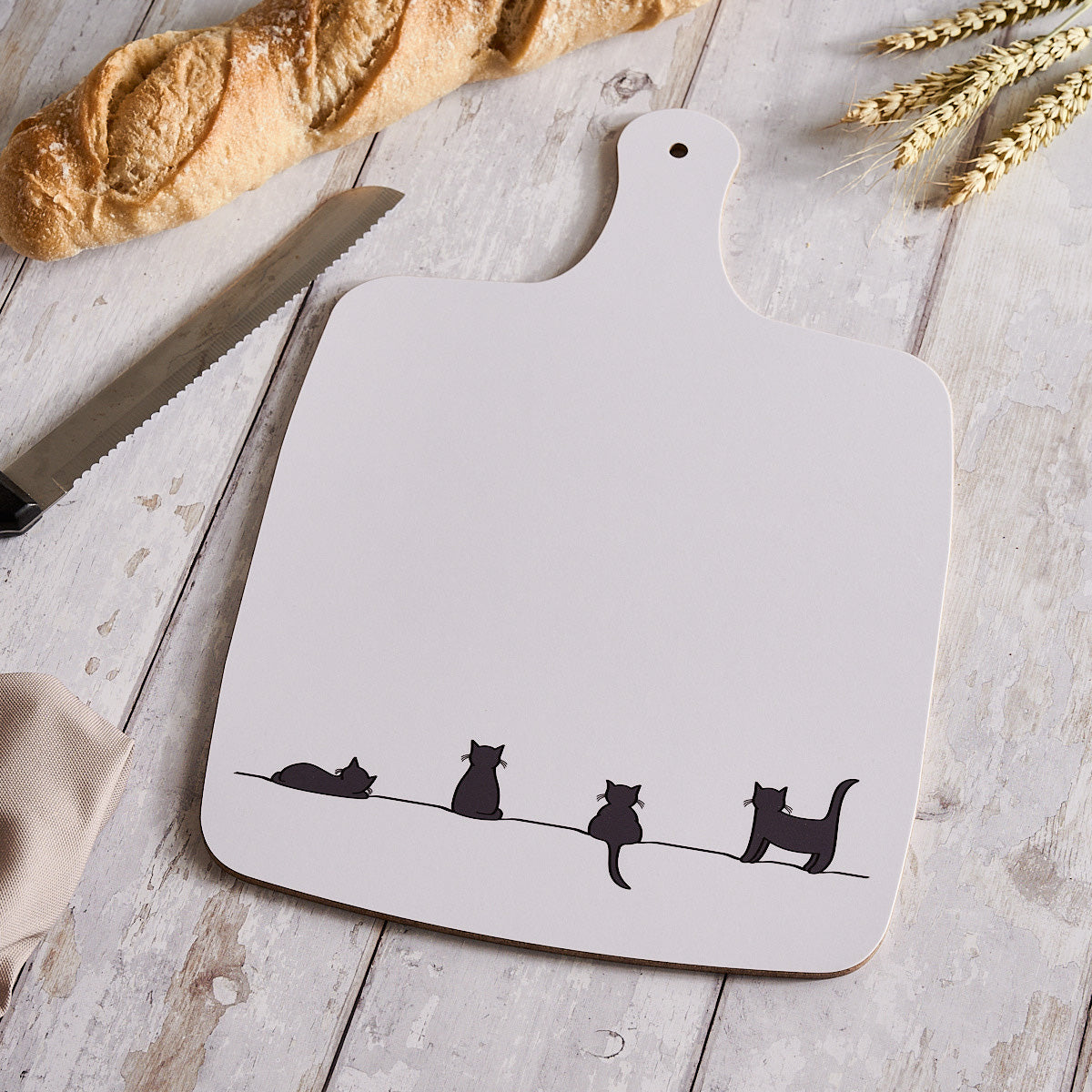 White cutting board with cat illustrations on a wooden surface with bread and wheat.