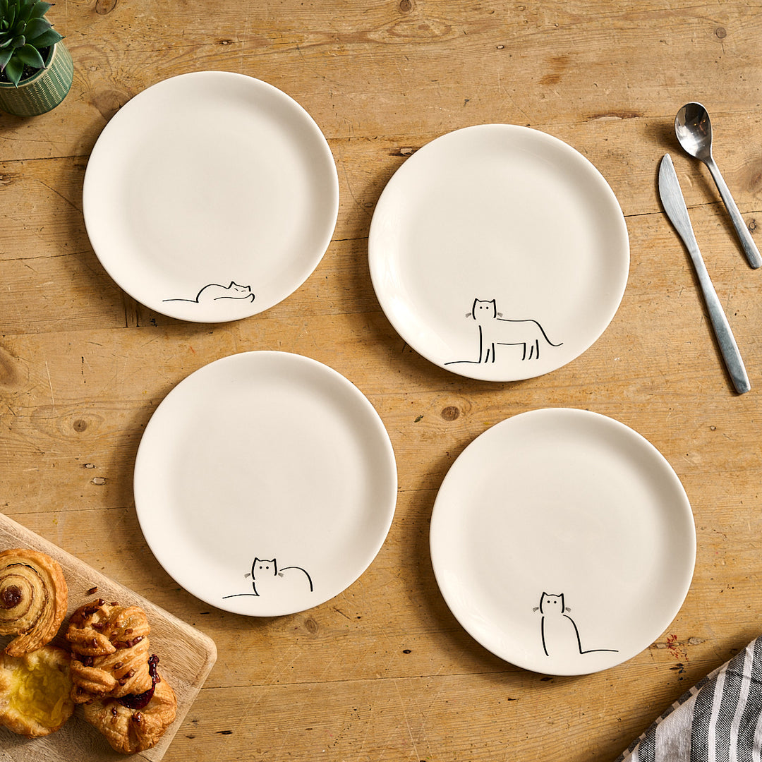 Four white plates with Jin Designs Cat Companions on a wooden table with utensils and pastries.