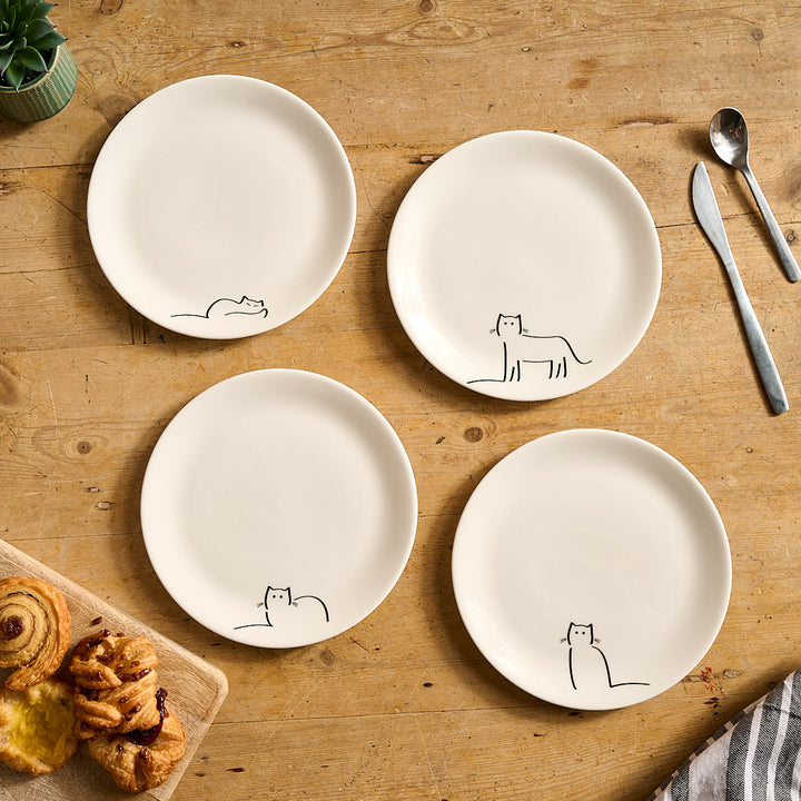 Four white plates with Jin Designs Cat Companions on a wooden table with utensils and pastries.