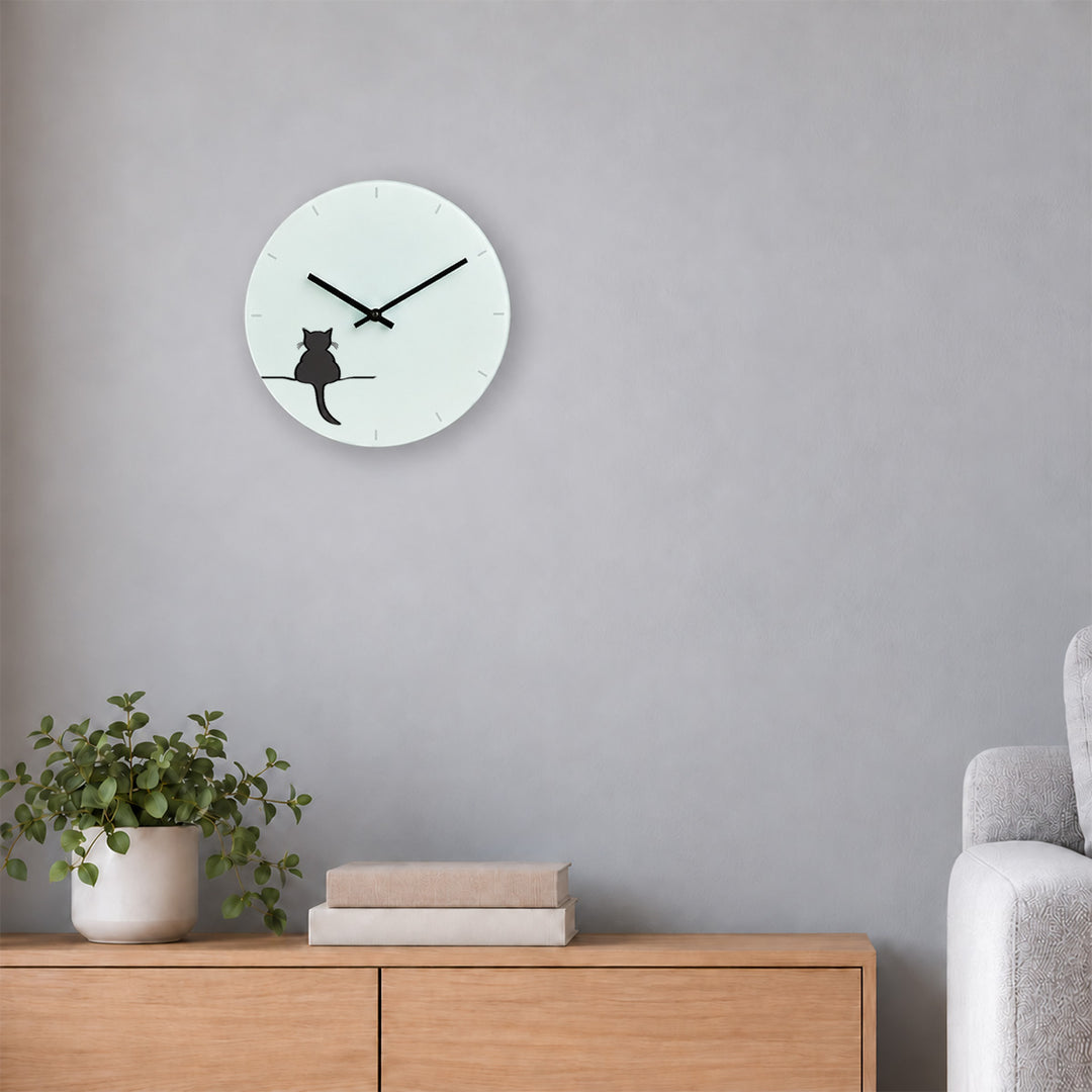 A minimalist round wall clock featuring Crouching Cat by Jin Designs on white face, mounted on a light grey wall above a wooden sideboard with a potted plant and books, next to a grey sofa.
