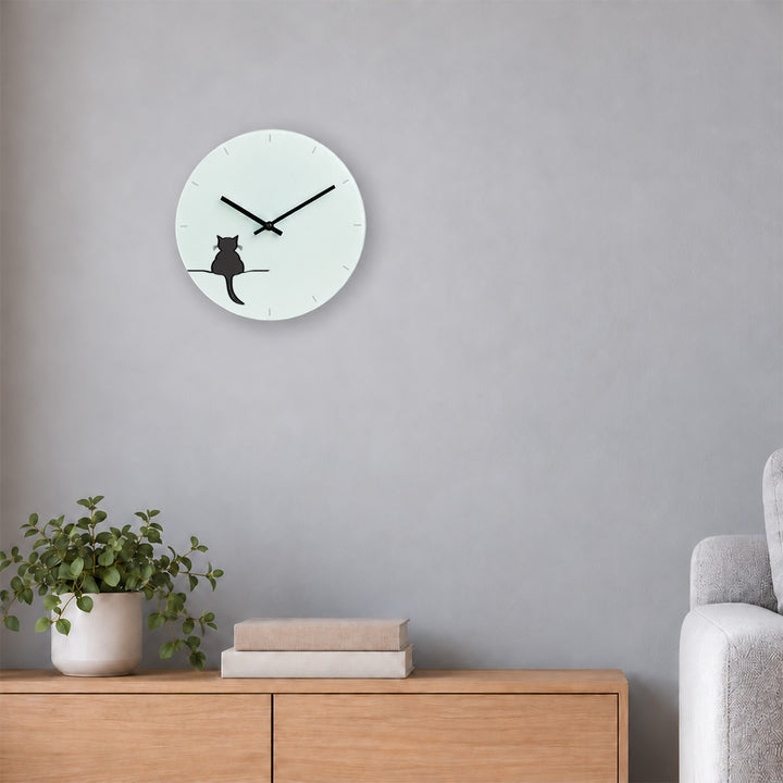 A minimalist round wall clock featuring Crouching Cat by Jin Designs on white face, mounted on a light grey wall above a wooden sideboard with a potted plant and books, next to a grey sofa.