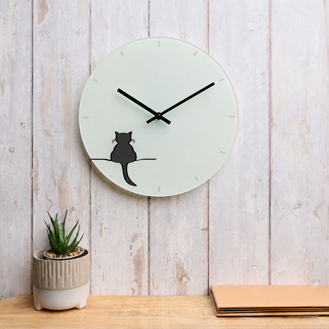 A minimalist round wall clock featuring Crouching Cat by Jin Designs. The clock is displayed on a light wood surface against a whitewashed wood plank wall, with a small potted succulent on the left and a stack of tan papers on the right.