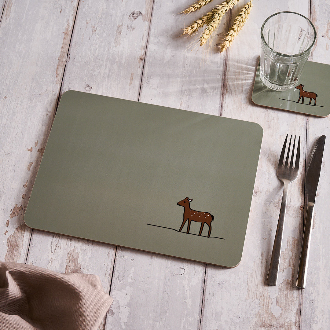 Sage Green placemat with a deer design on a wooden table with cutlery and a glass.
