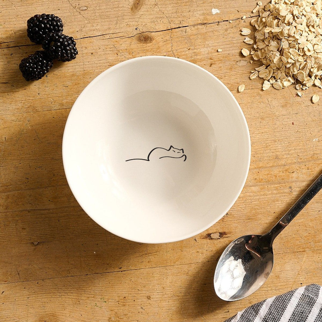 White bowl with Dreaming Cat on a wooden surface with blackberries, oats, and a spoon.