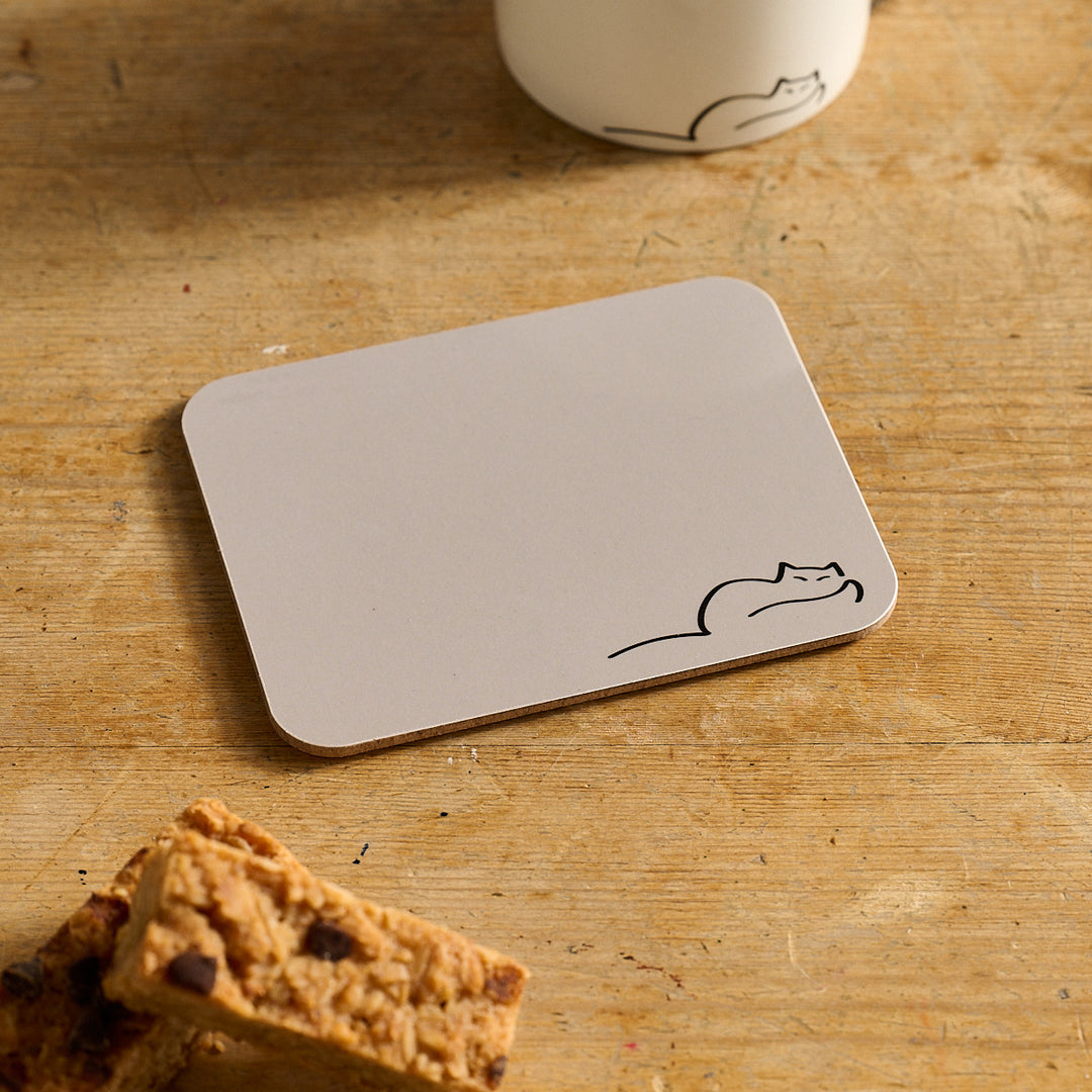 Dreaming Cat Coaster with a cat design on a wooden surface with a cup and snack.