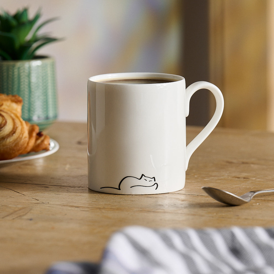 Dreaming Cat Mug with a cat illustration on a wooden table with a croissant and spoon.