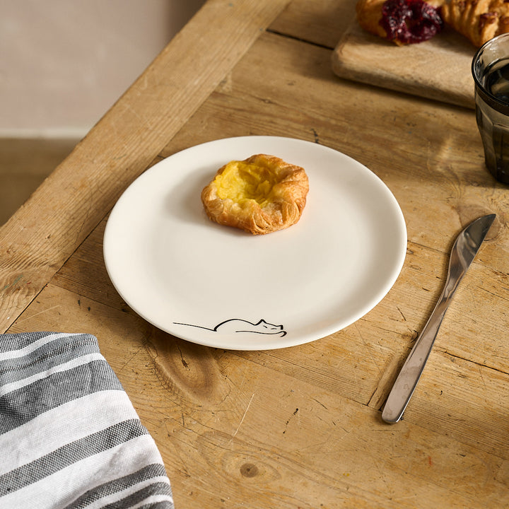 Dreaming Cat Side Plate with a croissant on a wooden table with a knife and striped cloth.