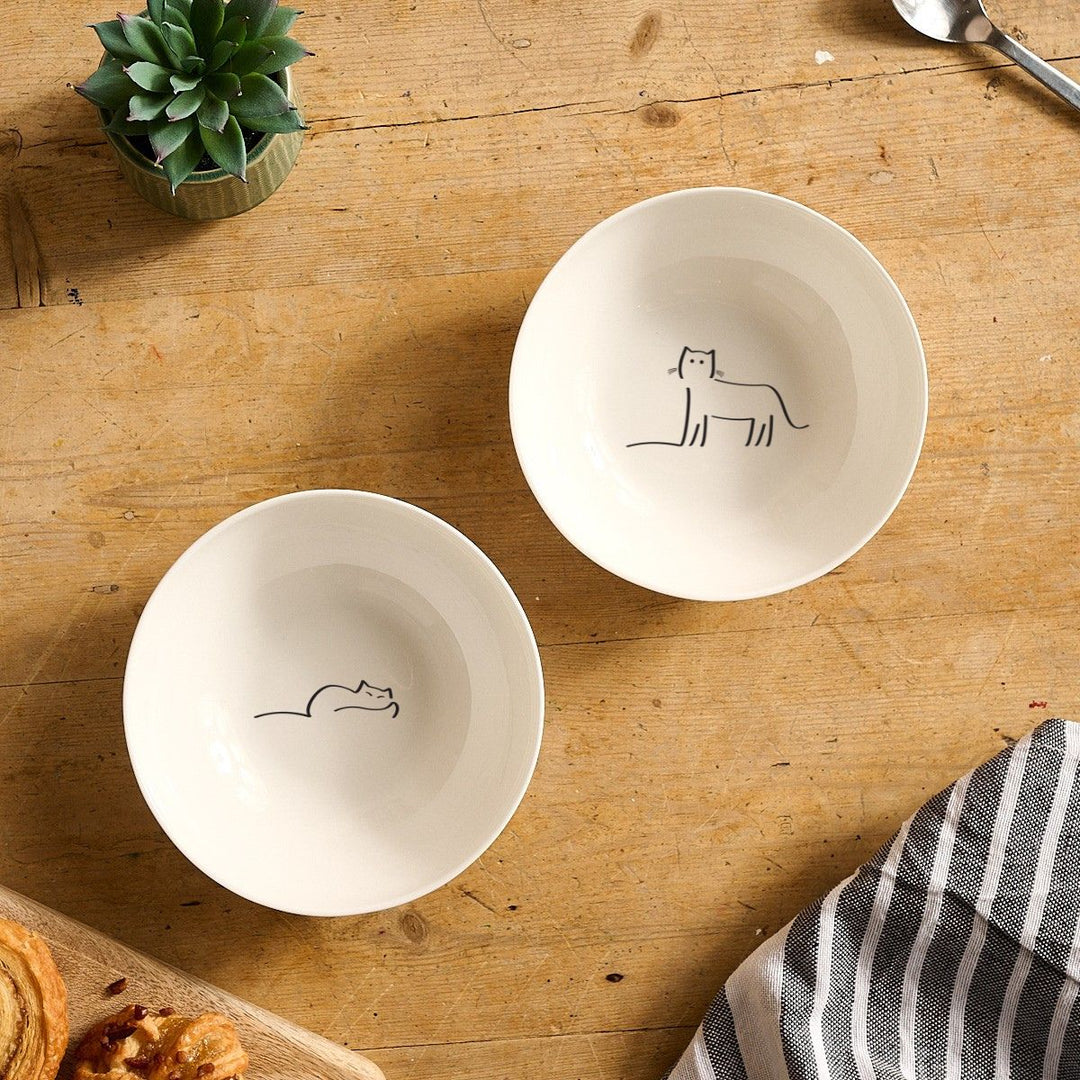 Two white bowls with Dreaming Cat and Watching Cat on a wooden table.