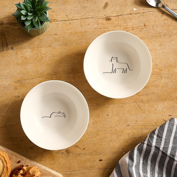Two white bowls with Dreaming Cat and Watching Cat on a wooden table.