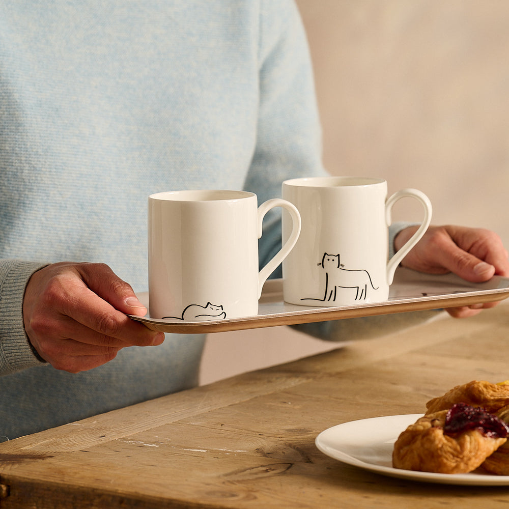 Two white mugs with cat illustrations on a wooden tray, held by a person, with a plate of pastries on a wooden table.