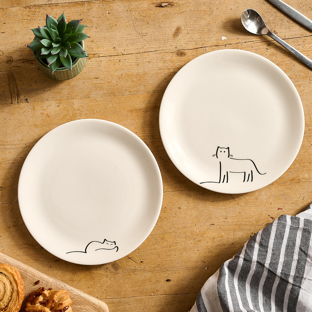 Dreaming Cat and Watching Cat  - Two white ceramic plates with cat illustrations on a wooden surface with a plant and utensils.