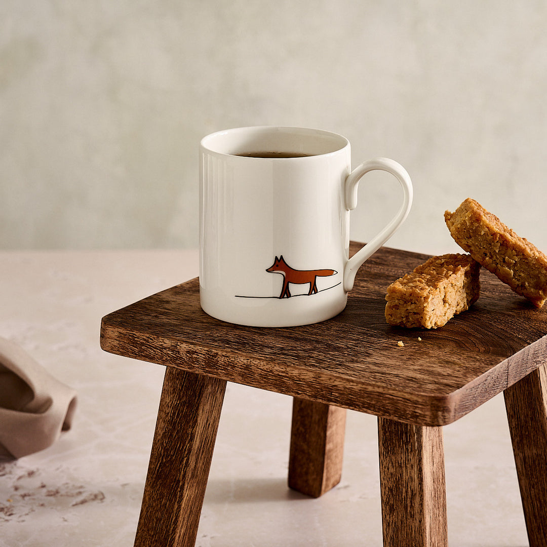 White mug with a fox design on a wooden stool with flapjacks