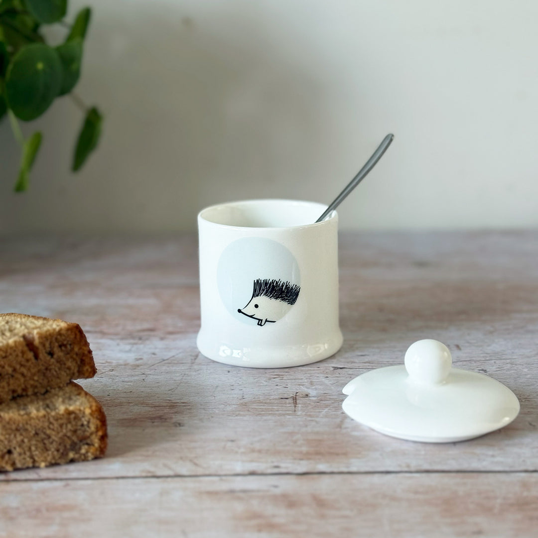White jam pot with a hedgehog design, spoon, and bread on a wooden surface.
