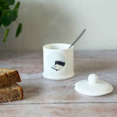 White jam pot with a hedgehog design, spoon, and bread on a wooden surface.