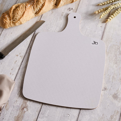 Grid-patterned cutting board with 'Jin Designs' branding on a wooden surface with bread and a knife.