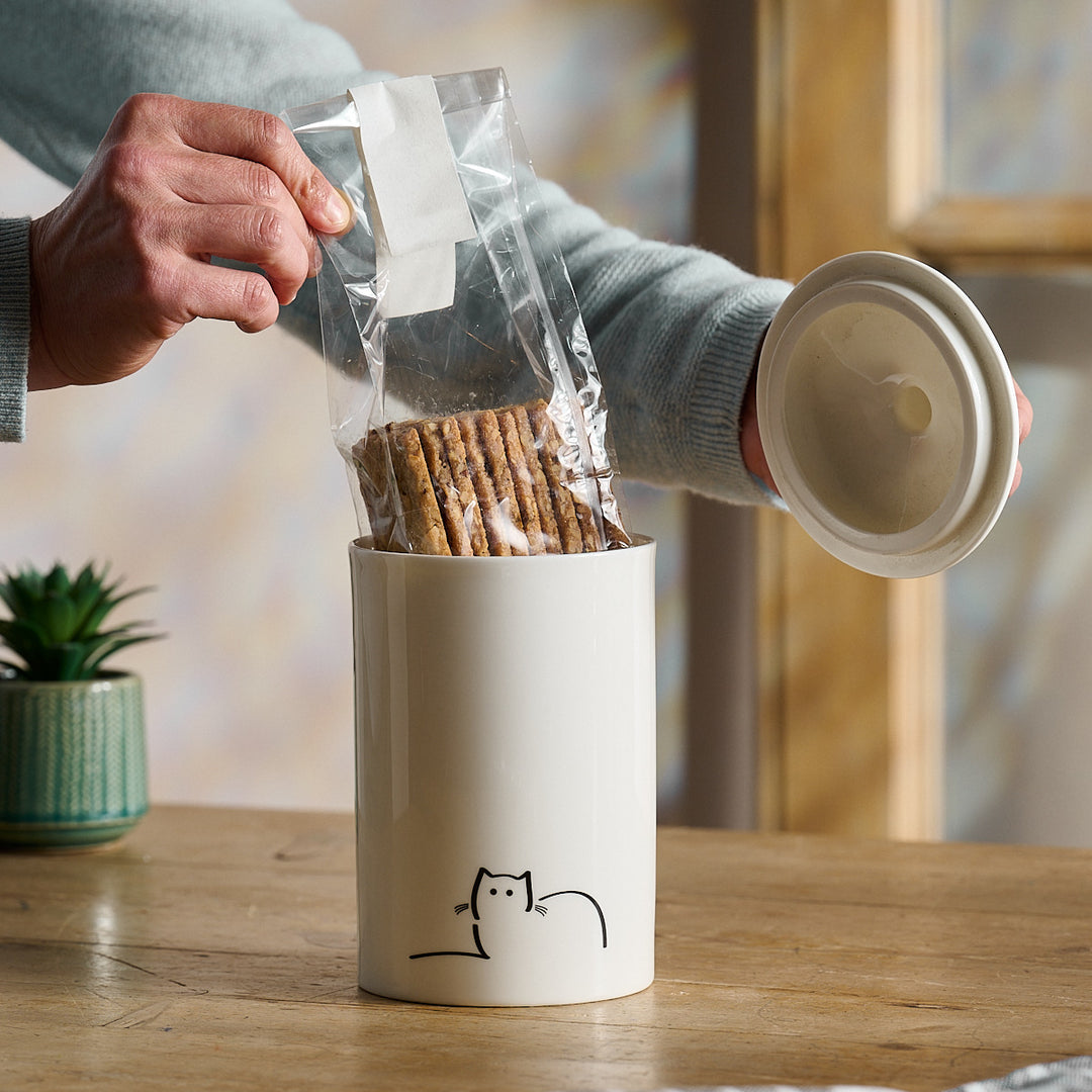 Person putting biscuits into a Loafing Cat Storage Jar with a cat design on a wooden table.