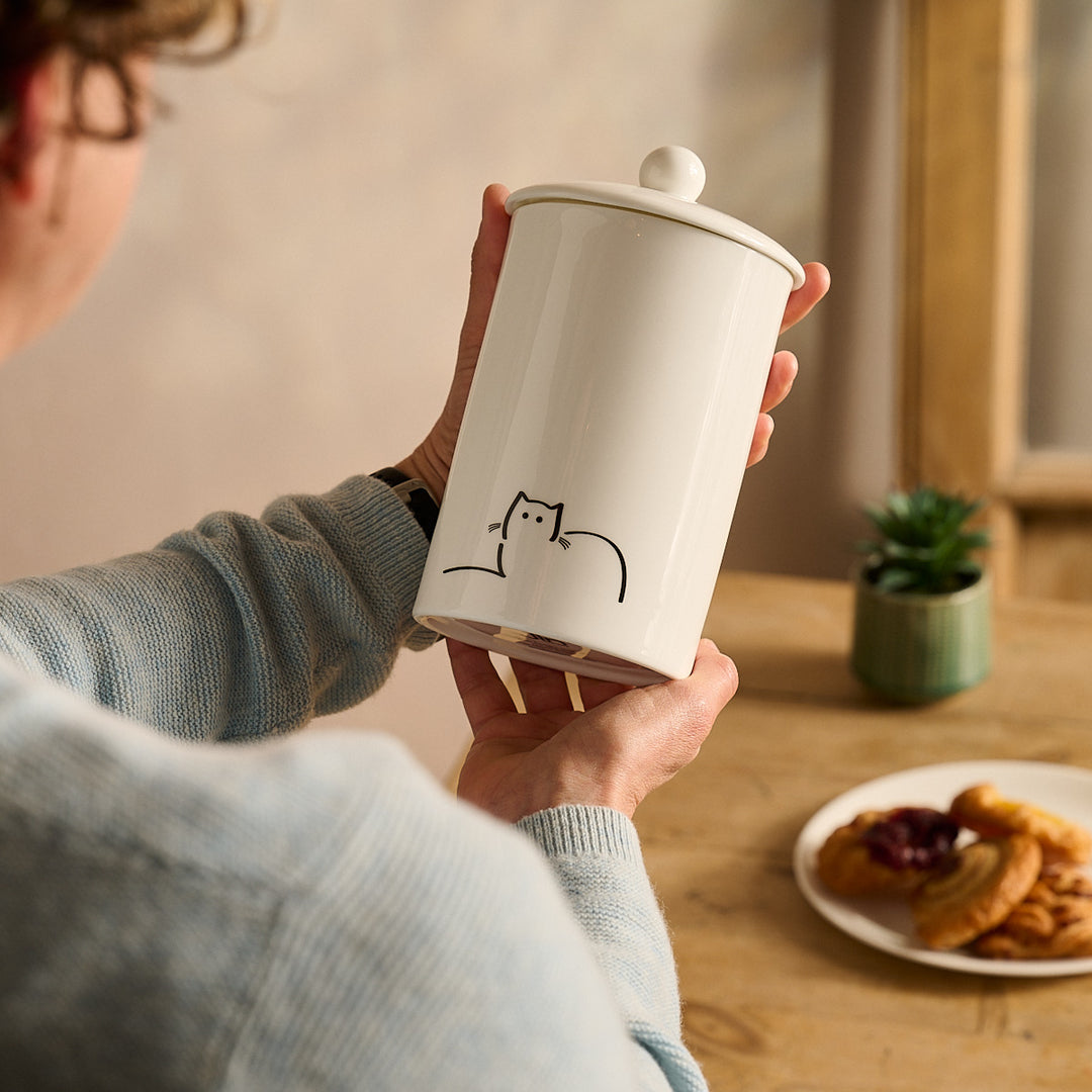 Person holding a Loafing Cat Storage Jar with a cat design on a wooden table with a plate of cookies and a plant.