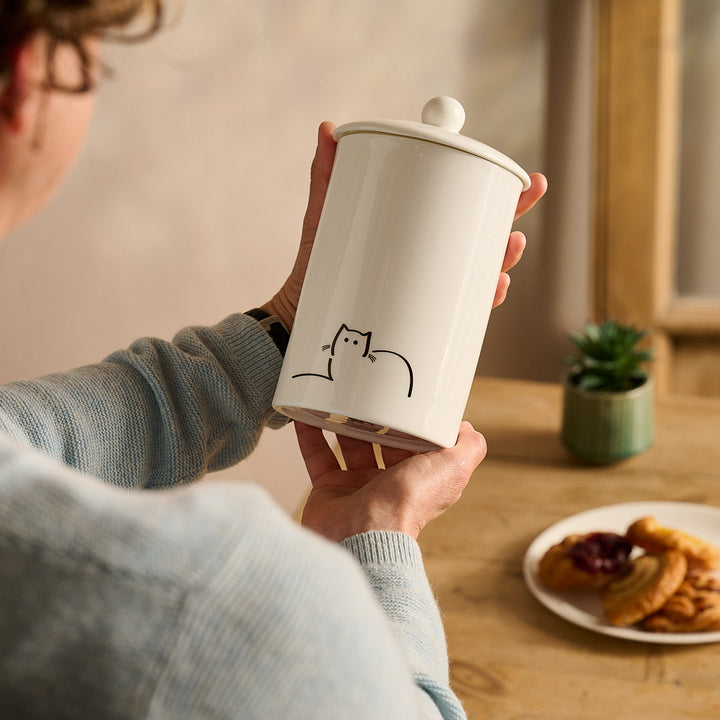 Person holding a Loafing Cat Storage Jar with a cat design on a wooden table with a plate of cookies and a plant.