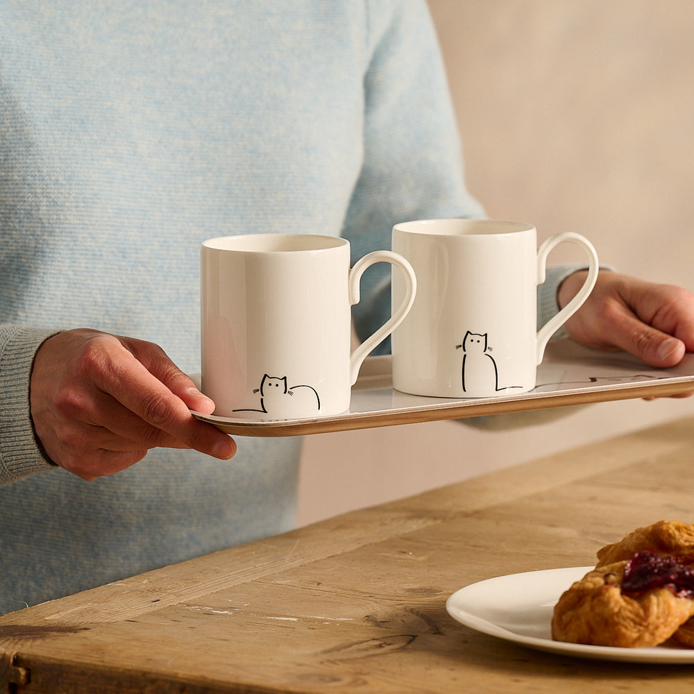 Loafing Cat and Waiting Cat - Two white mugs with cat illustrations on a tray, held by a person, with a plate of pastries on a wooden table.