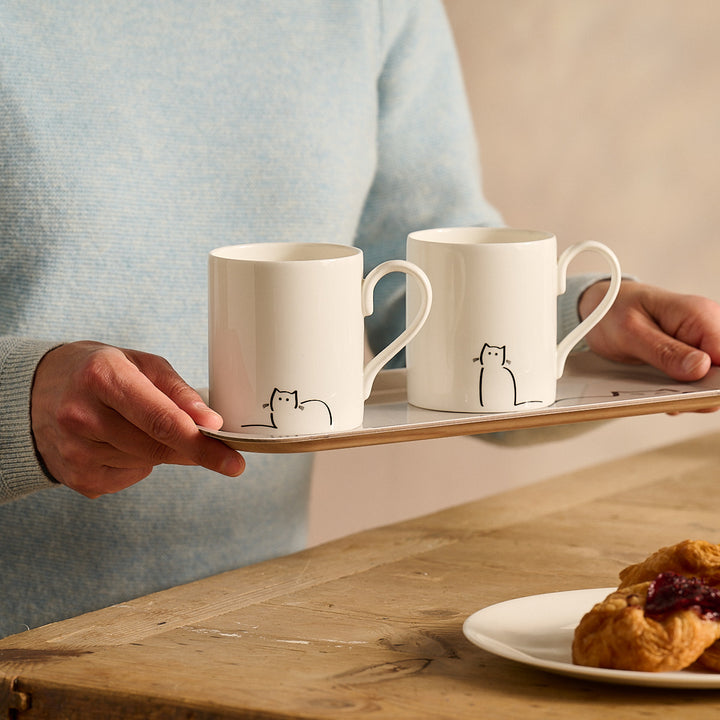 Loafing Cat and Waiting Cat - Two white mugs with cat illustrations on a tray, held by a person, with a plate of pastries on a wooden table.