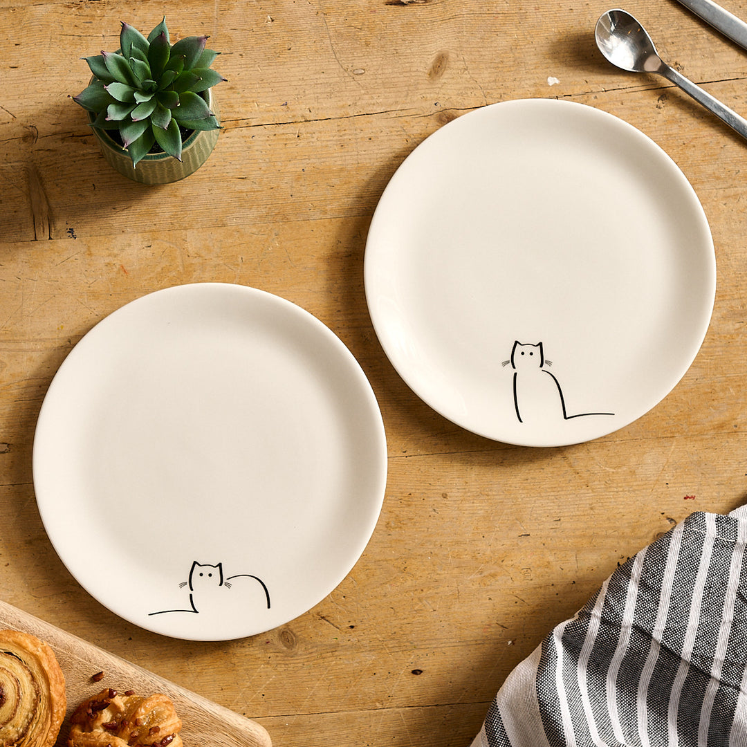 Two white plates with Loafing Cat and Waiting Cat on a wooden table with a plant and spoon in the background.