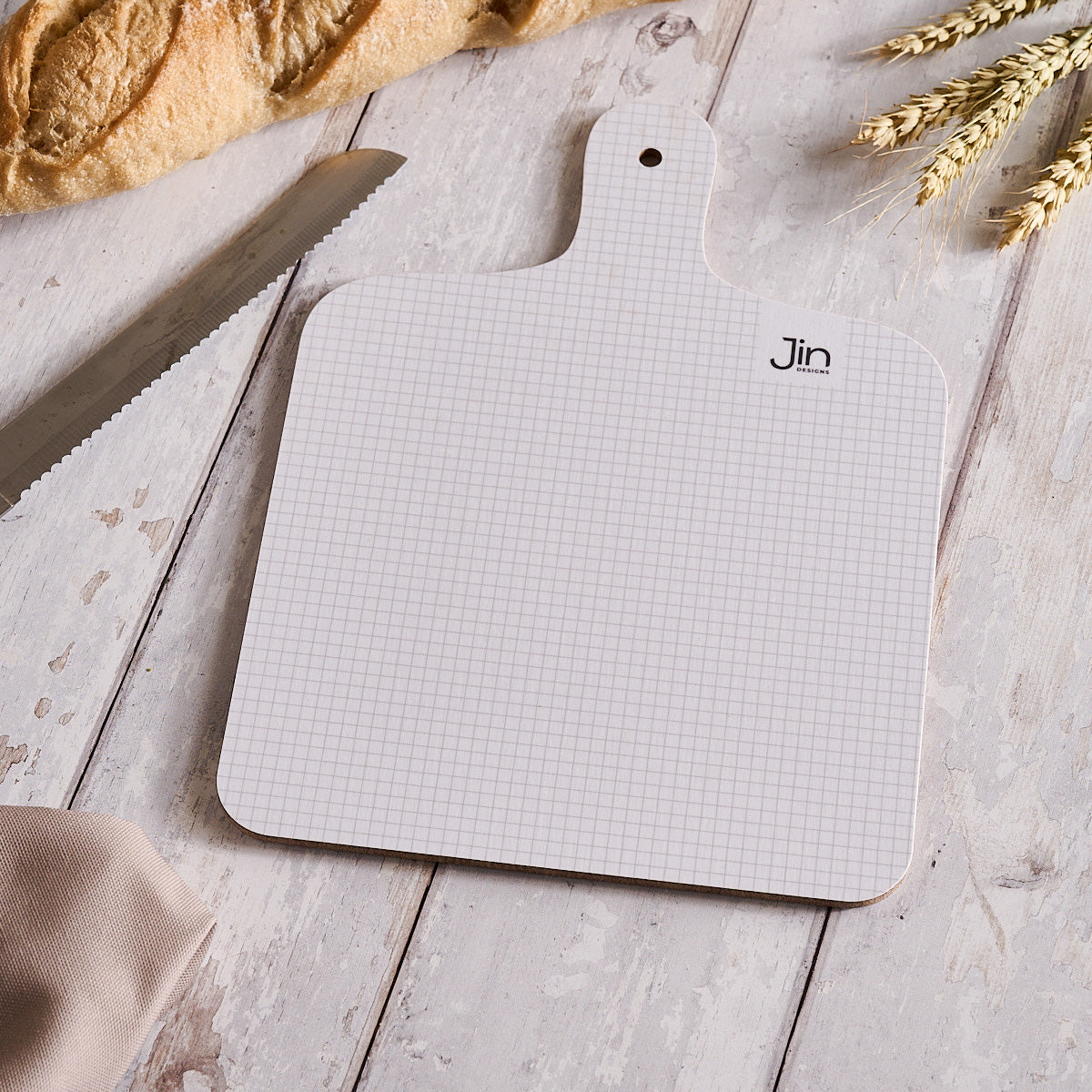 Grid-patterned cutting board with 'Jin Designs' branding on a wooden surface with bread and wheat.
