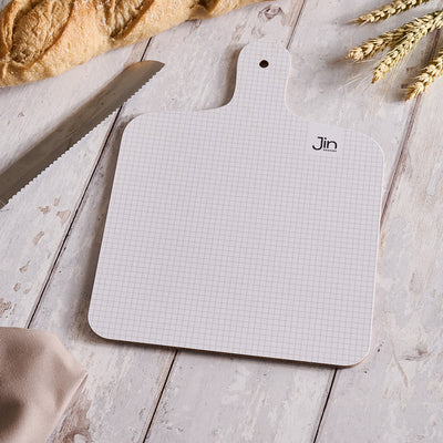 Grid-patterned cutting board with 'Jin Designs' branding on a wooden surface with bread and wheat.