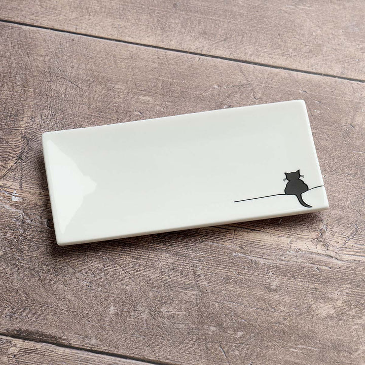 A rectangular glossy white ceramic dish with a small black cat silhouette sitting on a thin line near the bottom right, displayed on a rustic brown wooden surface.