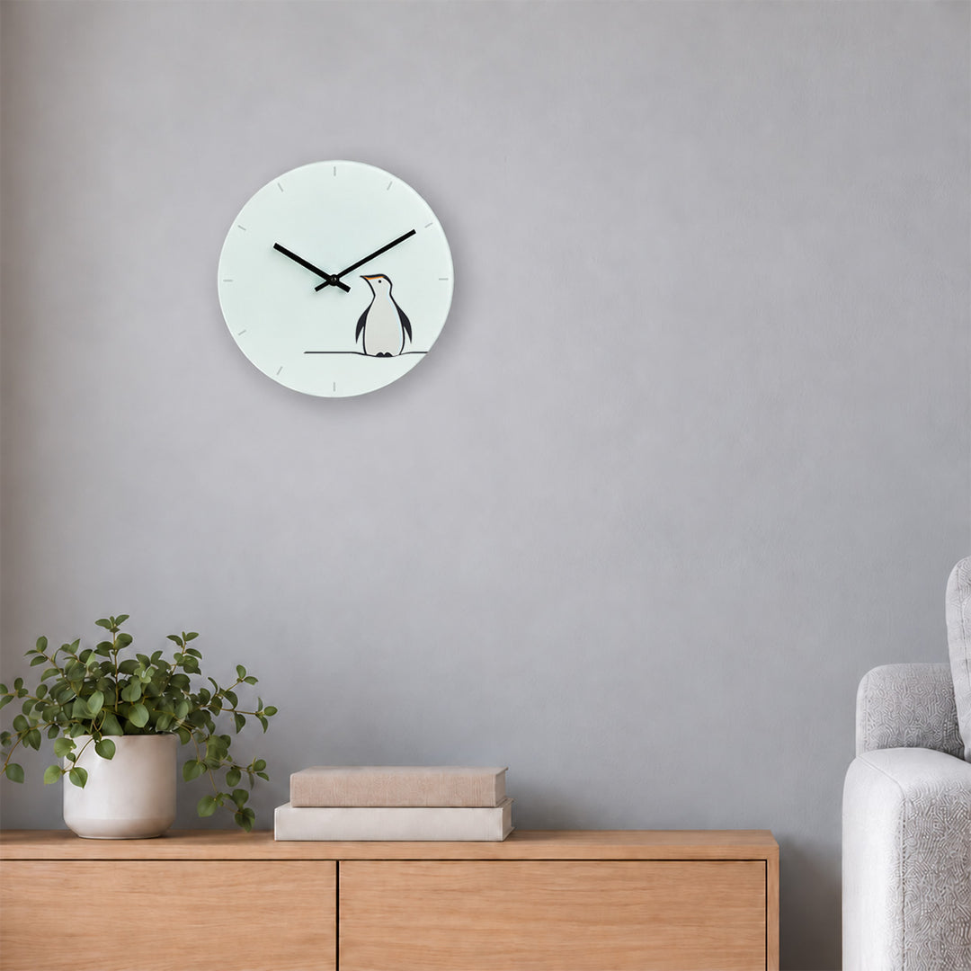 A minimalist white round wall clock featuring Penguin by Jin Designs mounted on a wall above a wooden console table with a potted plant and books.