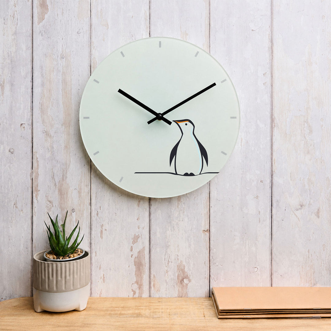 A minimalist round wall clock featuring Penguin by Jin Designs. The clock is mounted on a light wood-paneled wall above a wooden surface, with a small potted succulent on the left and stacked papers on the right.