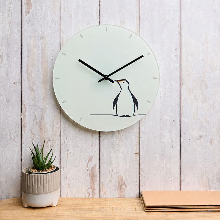 A minimalist round wall clock featuring Penguin by Jin Designs. The clock is mounted on a light wood-paneled wall above a wooden surface, with a small potted succulent on the left and stacked papers on the right.