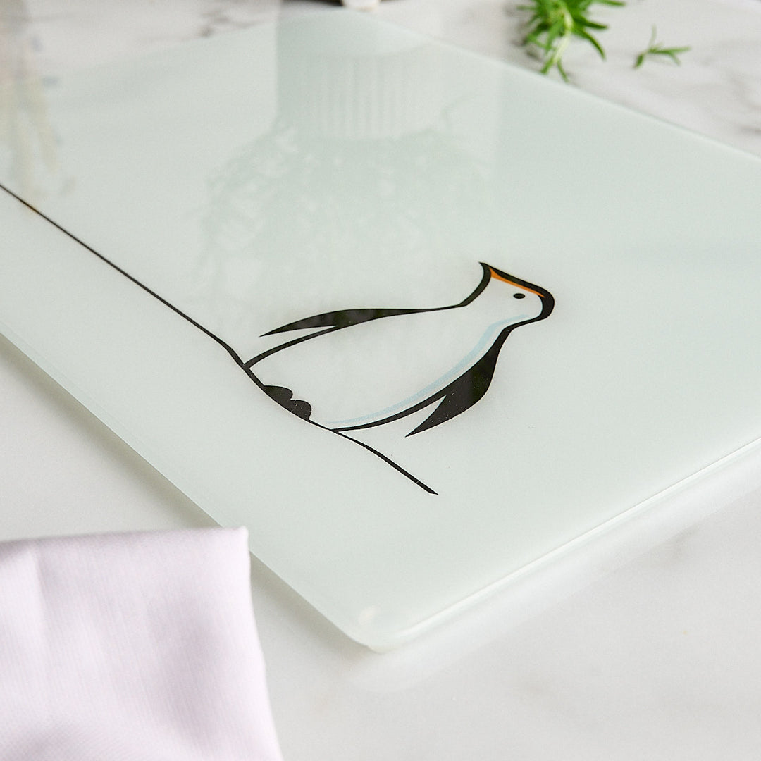 A frosted glass worktop saver or serving board featuring a minimalist black penguin illustration, styled on a white marble surface with a white napkin and green leaves in the background.