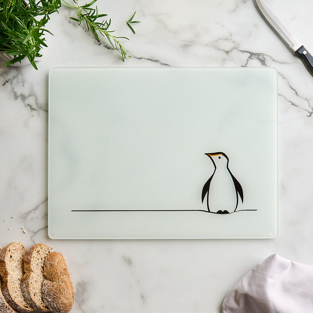 A frosted white rectangular worktop saver or cutting board with a minimalist black line drawing of a penguin, styled on a white marble surface with bread, herbs, and a knife.