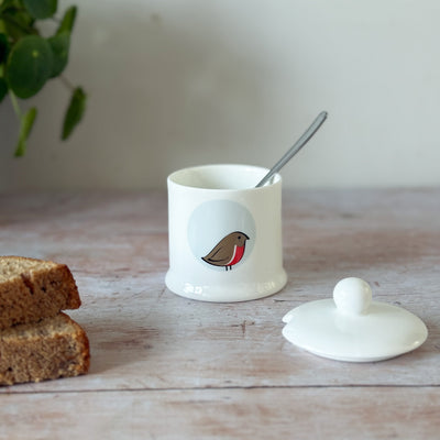Small white fine bone china jam pot with a robin bird design on a wooden surface with cake and a plant.
