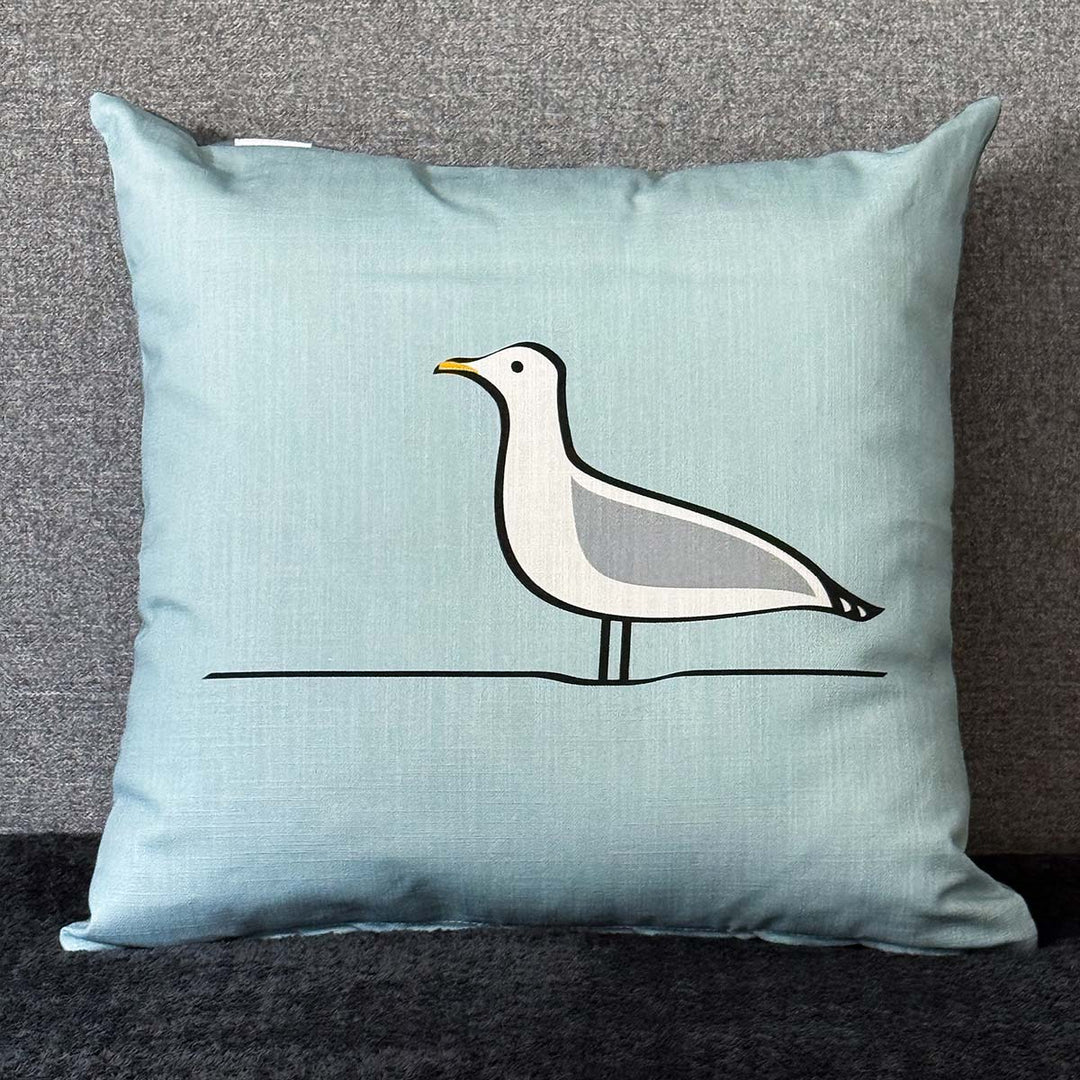 A square light blue cushion featuring a minimalist Seagull design in white, grey, black, and yellow, set against a textured gray background.