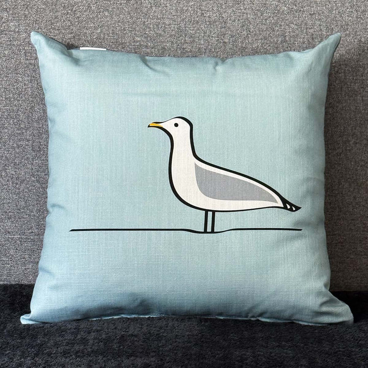 A square light blue cushion featuring a minimalist Seagull design in white, grey, black, and yellow, set against a textured gray background.