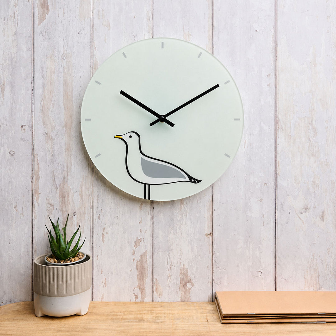 A minimalist round glass wall clock featuring Seagull by Jin Designs, displayed on a light wooden wall against a whitewashed wood plank wall, next to a small potted succulent and stacked papers.