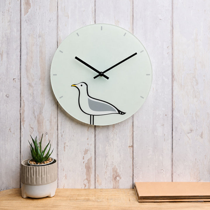 A minimalist round glass wall clock featuring Seagull by Jin Designs, displayed on a light wooden wall against a whitewashed wood plank wall, next to a small potted succulent and stacked papers.