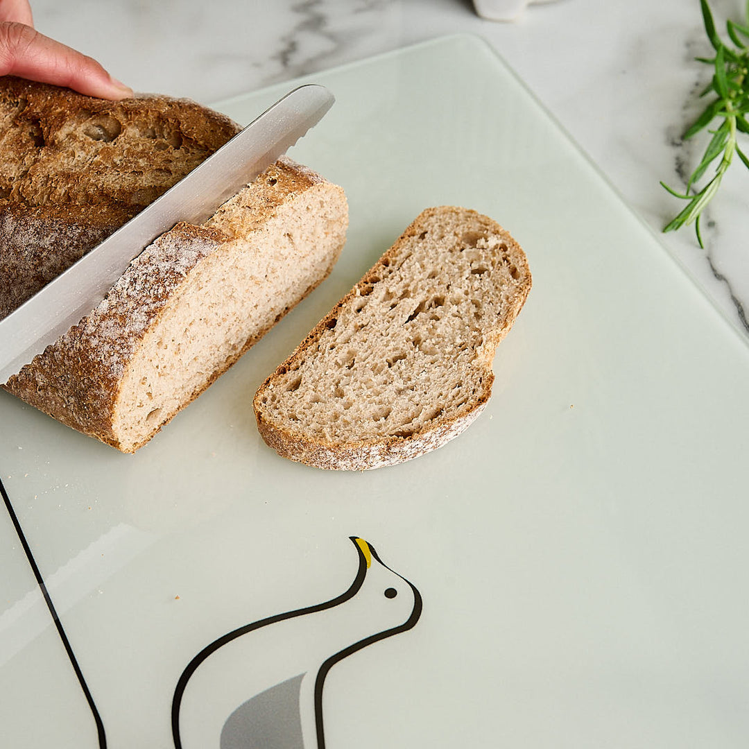 A hand is slicing a rustic loaf of bread on a white glass worktop saver or cutting board with a Seagull design by Jin Designs. A slice of bread rests beside the board on a light marble surface with a small green herb sprig in the corner.