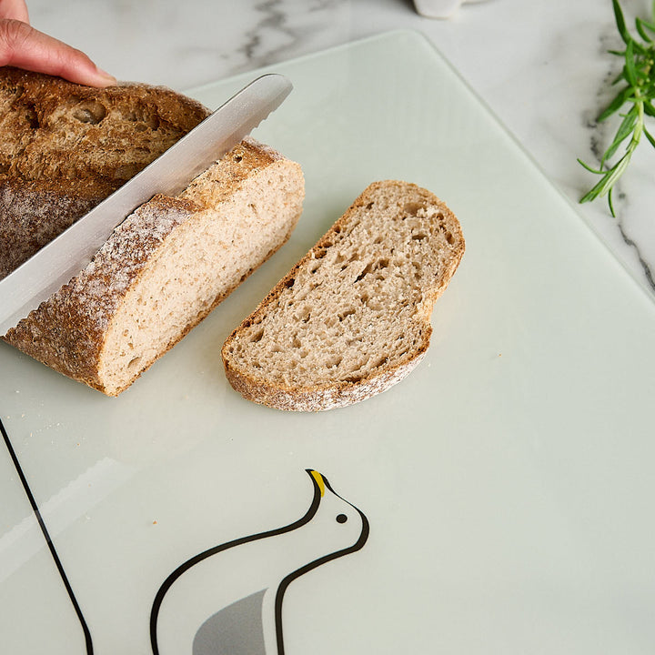 A hand is slicing a rustic loaf of bread on a white glass worktop saver or cutting board with a Seagull design by Jin Designs. A slice of bread rests beside the board on a light marble surface with a small green herb sprig in the corner.