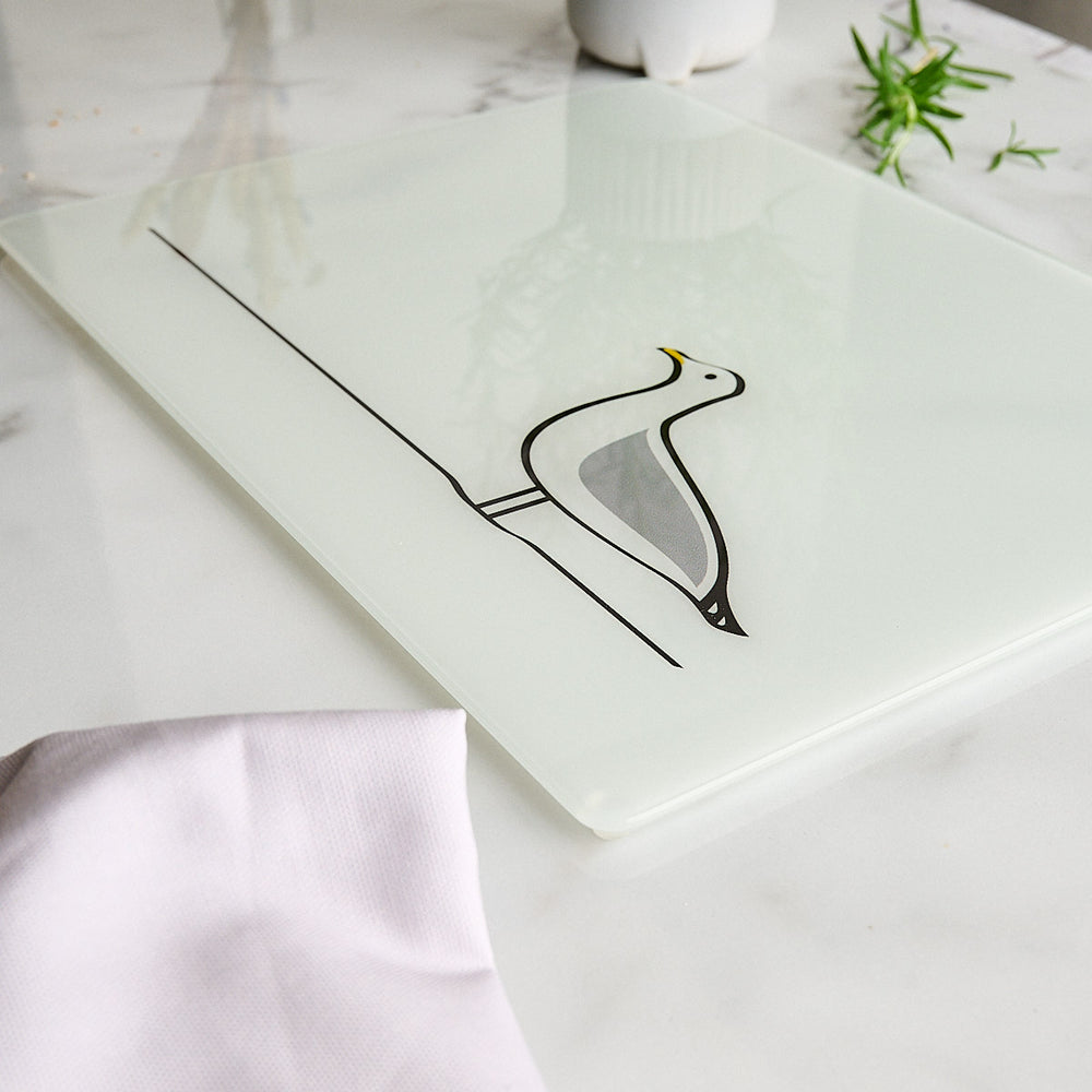 A rectangle white glass worktop saver or chopping board featuring a minimalist Seagull design by Jin Designs, styled on a white marble surface with a light pink cloth and green herbs.