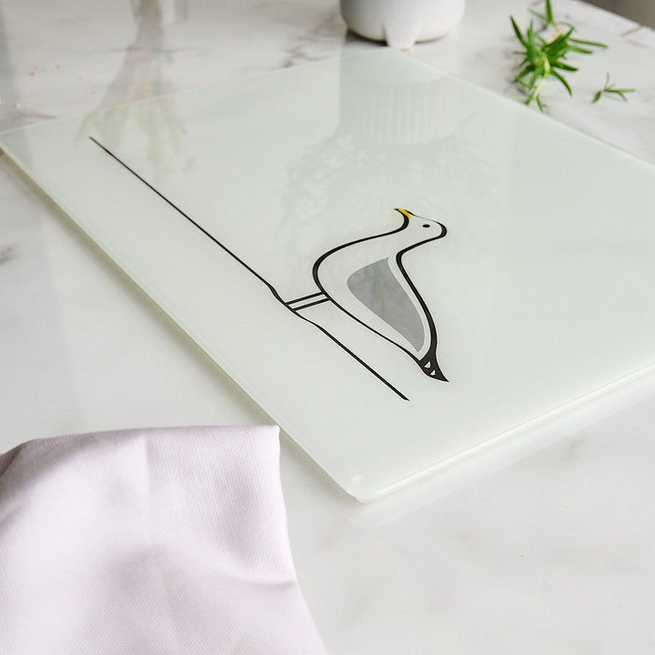A rectangle white glass worktop saver or chopping board featuring a minimalist Seagull design by Jin Designs, styled on a white marble surface with a light pink cloth and green herbs.