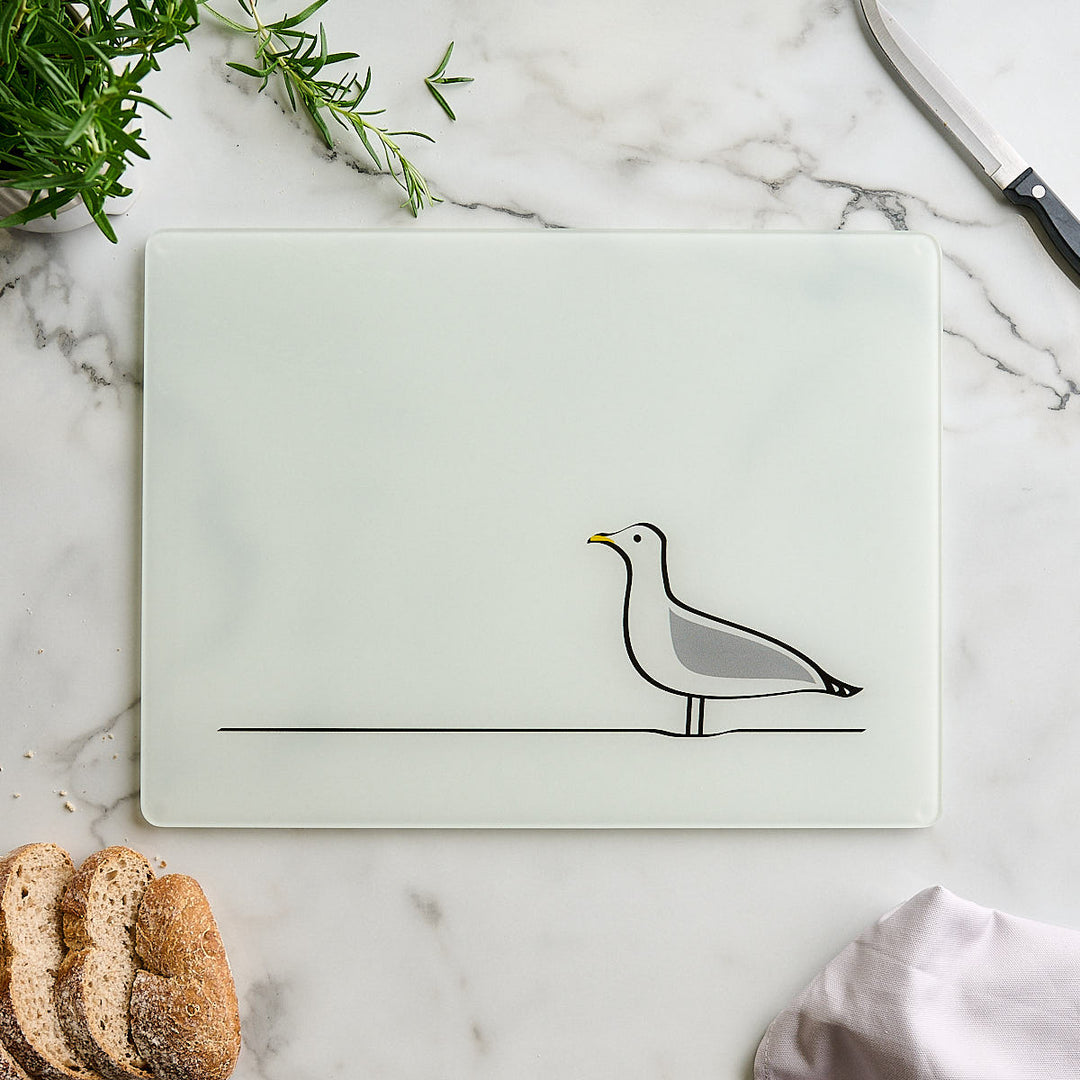 A rectangular white glass worktop saver with rounded corners, featuring Jin Designs Seagull. The cutting board is styled in a flat-lay on a white marble surface, surrounded by a knife, bread, and green herbs.