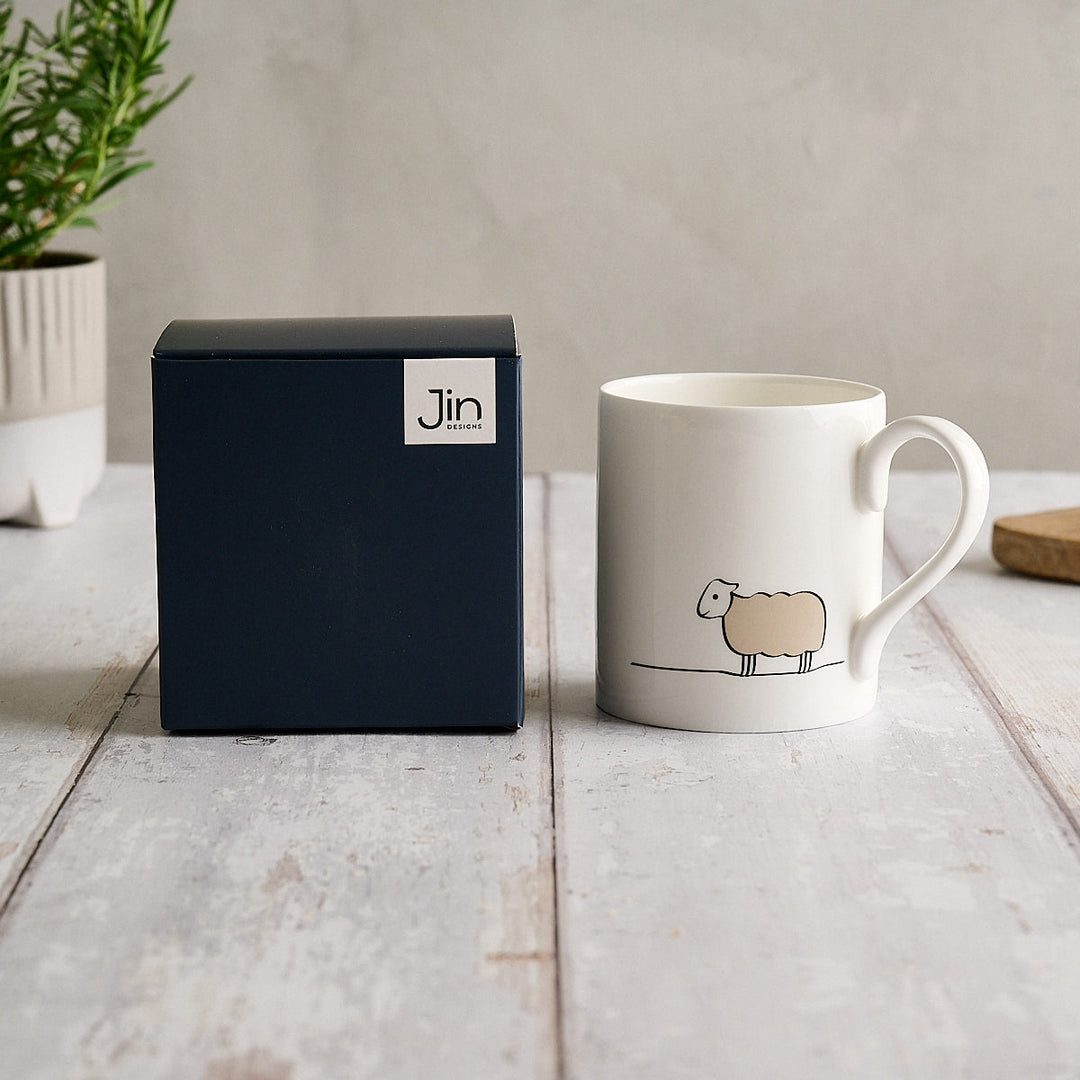 A Jin Designs white fine bone china Sheep Mug next to its dark navy blue Jin Designs Gift box on a light wooden tabletop.