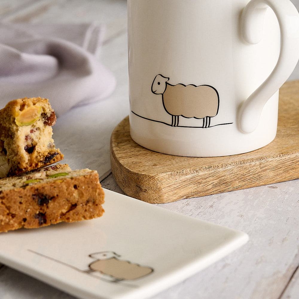 A white fine bone china mug with a minimalist sheep illustration sits on a round wooden coaster, next to sliced fruit on a white plate with a matching illustration. The scene is styled on a light gray wooden tabletop with a soft lavender cloth in the background.