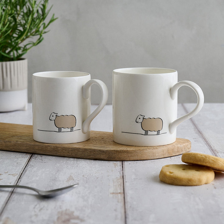 Two white ceramic mugs with a minimalist sheep illustration, displayed on a wooden board on a light gray rustic table with cookies and a plant in the background.