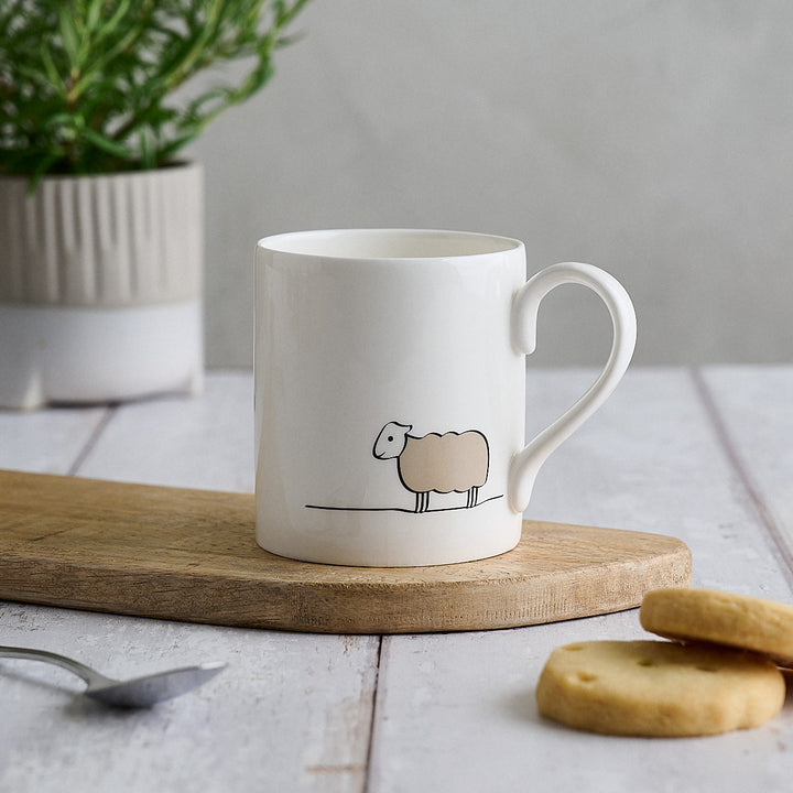 A white fine bone china mug with a minimalist sheep illustration, resting on a wooden board on a light tabletop with biscuits and a potted plant in the background.